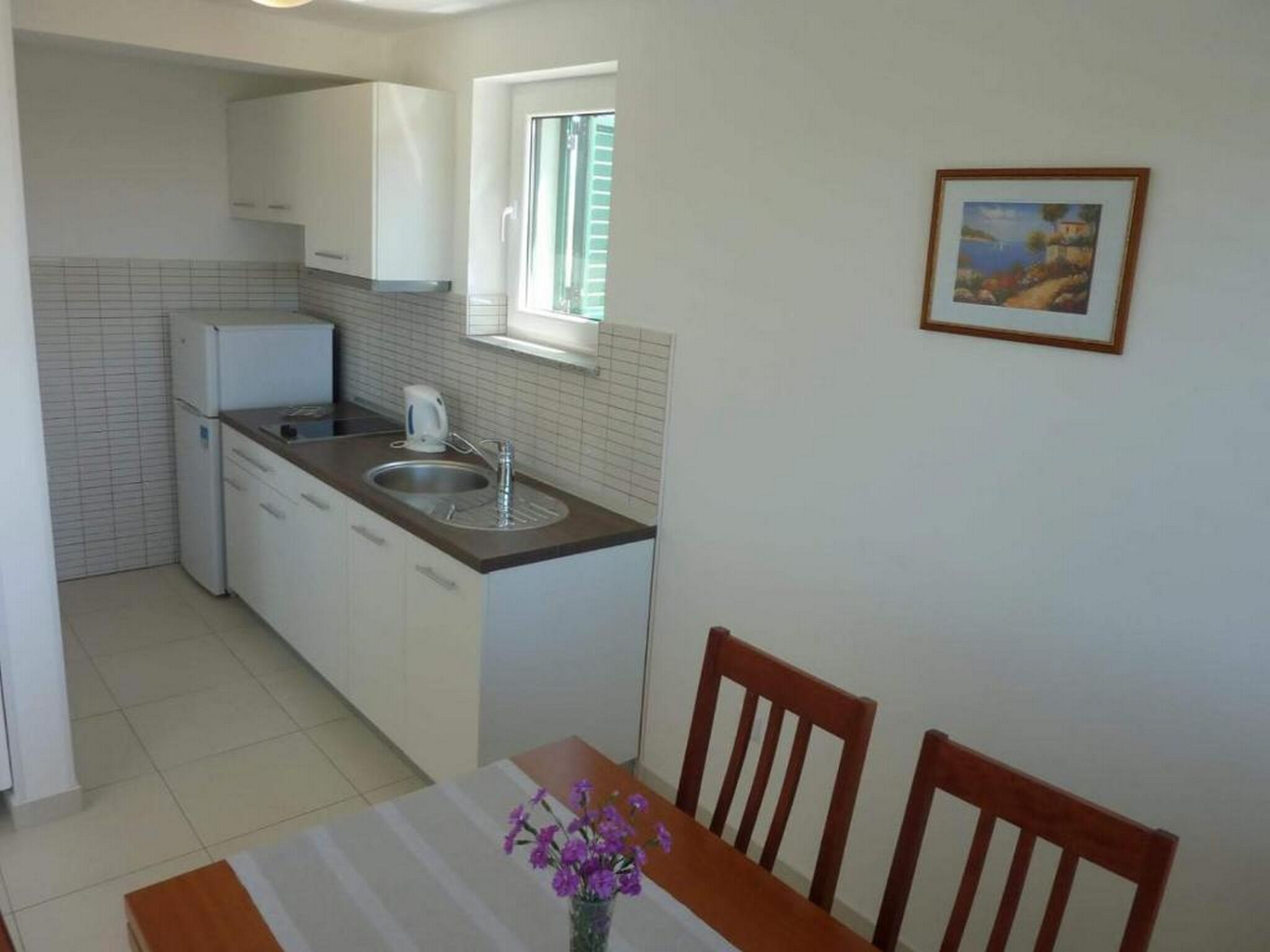 A compact kitchen with white cabinets, a countertop, a sink, and a window. There is a dining table with chairs and a vase of flowers.