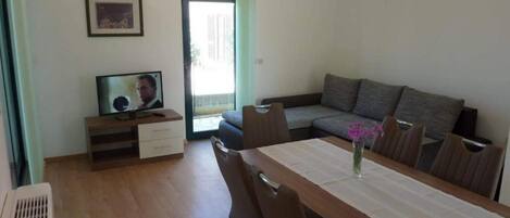 A living room with a sofa, a TV on a wooden stand, a dining table with chairs, and a window with curtains.