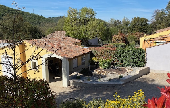 Un bâtiment jaune avec un toit en tuiles, entouré d’un jardin où poussent différentes espèces de plantes et où se trouve un petit étang.