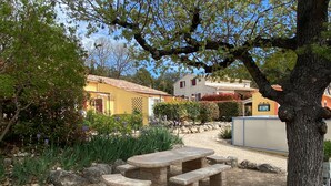 Ein Steintisch mit Bänken in einem Gartenbereich, umgeben von Pflanzen und mit einem Gebäude im Hintergrund.