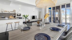 Een moderne eetruimte met een lange houten tafel voor zes personen, een grote geweven lamp boven de tafel en uitzicht op de omgeving door grote ramen.
