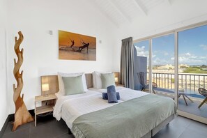 Ein Hotelzimmer mit einem großen Bett, einem Nachttisch, einer Holzskulptur, einem Gemälde mit Strandmotiv und einem Balkon mit Blick auf den Strand.