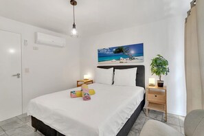 Une chambre d’hôtel comprenant un lit, des tables de chevet, une petite table, un climatiseur fixé au mur, un luminaire au plafond, un tableau représentant une scène de plage et une plante en pot.