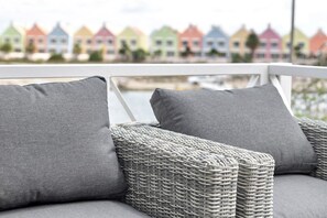 Un canapé en osier avec des coussins gris et une balustrade blanche au premier plan, et des maisons colorées en arrière-plan.