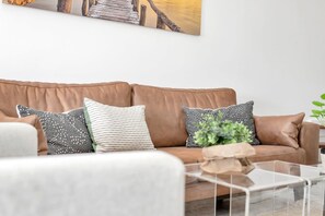 A brown leather sofa with patterned cushions, a white armchair, and a glass coffee table with a plant.