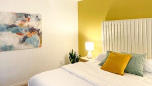 Un dormitorio moderno con cama, mesita de noche con lámpara, ventilador de techo y un cuadro abstracto colgado en la pared.