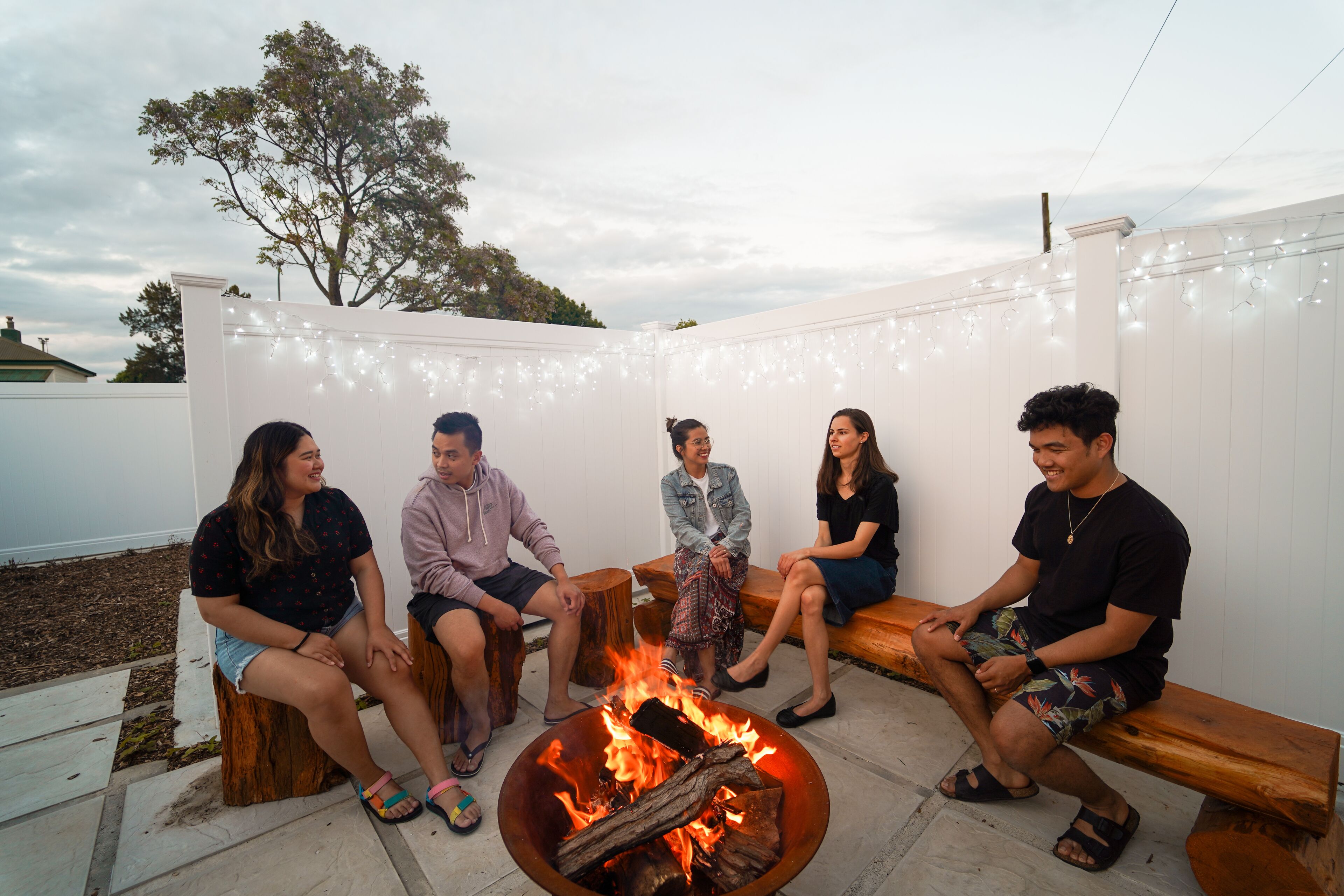 A group of people sitting around a fire pit in an outdoor patio area with string lights.