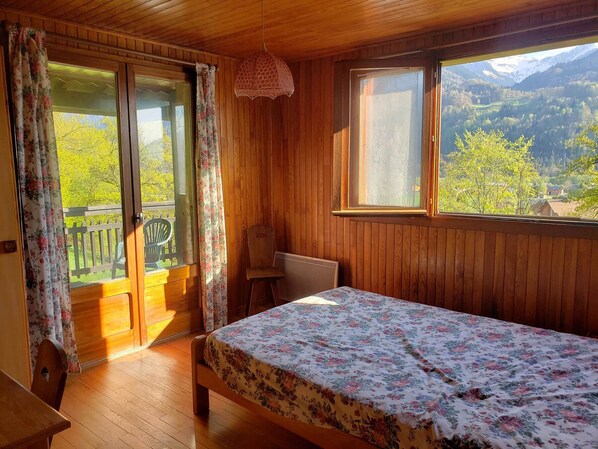 A wooden cabin room with a bed, a chair, and a view of mountains through a window.