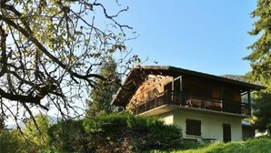 Une maison à deux étages avec un balcon, entourée de verdure et d’arbres.