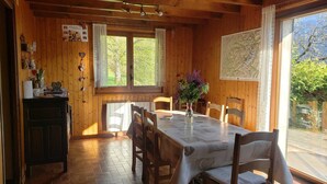 Une salle à manger en bois avec une table dressée pour quatre personnes, une fenêtre avec des rideaux et une vue sur la verdure à l’extérieur.