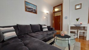 A living room with a dark grey sofa, a glass coffee table, and a painting of boats on the wall. - Apartment Marina Obod (Konavle)