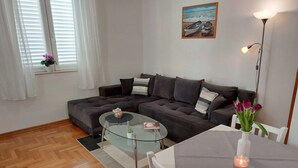 A modern living room with a sectional sofa, a glass coffee table, a dining table with chairs, a small potted plant, a painting on the wall, and a floor lamp. - Apartment Marina Obod (Konavle)