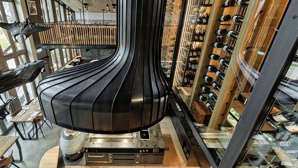 Een modern interieur met een centrale zwarte constructie, houten planken met wijnflessen en een eethoek met tafels en stoelen.
