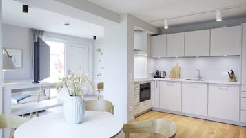 A modern kitchen with white cabinets, a built-in oven, and a sink. There is a dining area with a round table and chairs, and a vase with dried flowers on the table.