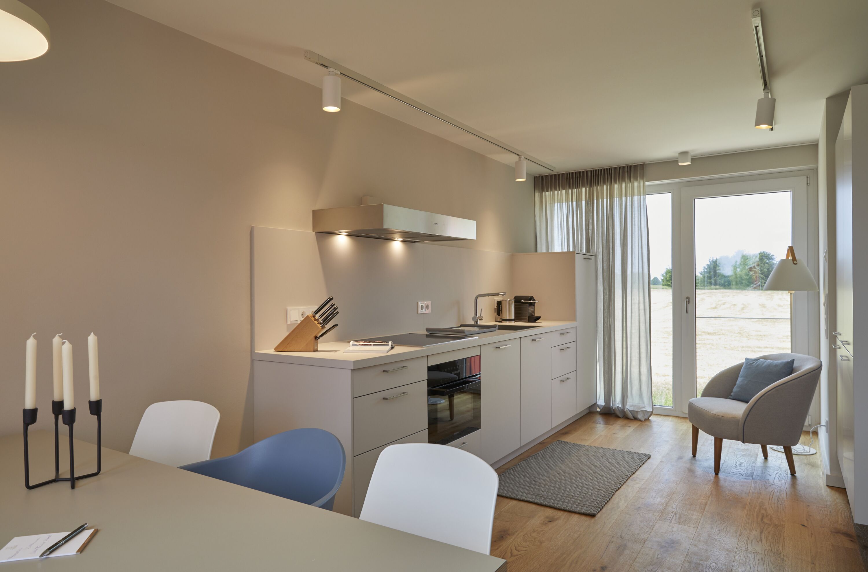 A modern kitchen with a dining area, featuring a built-in oven, a coffee maker, and a window with curtains.