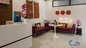 A reception area with a white reception desk, a purple sofa, a small side table, and decorative wall art including clocks and woven panels. - Aqua Inn (Kinshasa)
