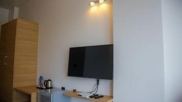 Une pièce compacte dotée d’un téléviseur à écran plat fixé au mur, d’une petite kitchenette avec un réfrigérateur, d’un meuble en bois et d’une étagère avec une bouilloire et des télécommandes.