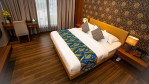A hotel room with a large bed, wooden floor, a desk with a chair, a nightstand with a lamp, and a patterned wallpaper. - Aqua Inn (Kinshasa)