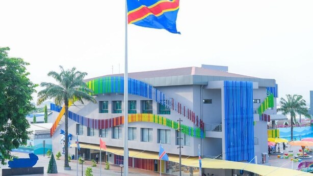 A modern building with a flagpole, palm trees, and a colorful facade. - Aqua Inn (Kinshasa)