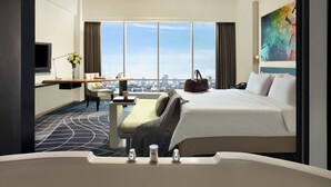 A hotel room with a large bed, a desk with a chair, a view of the city through large windows, and a bathtub. - ARTOTEL Grand Prima Surabaya (Surabaya)