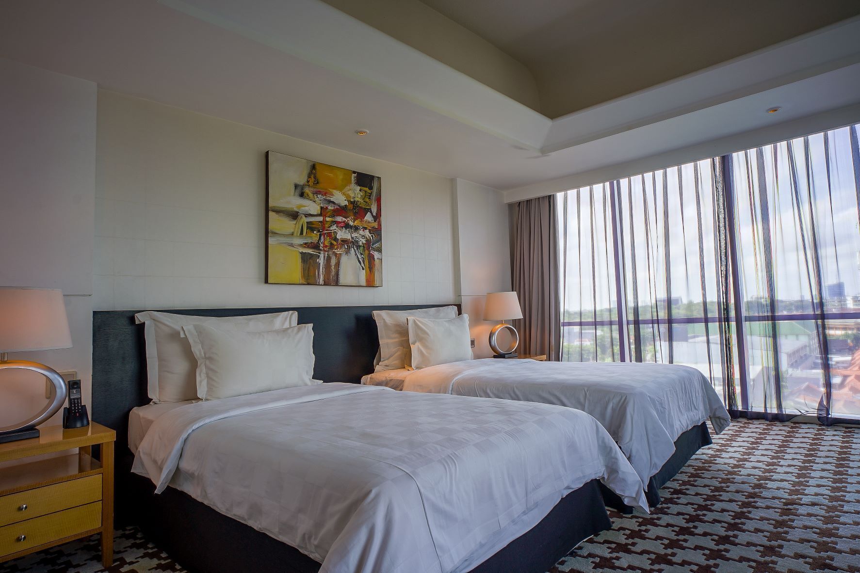 A hotel room with two beds, a painting on the wall, a window with curtains, and a patterned carpet.