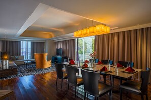 Ein moderner Essbereich mit einem langen Tisch, gedeckt für sechs Personen, umgeben von dunklen Stühlen. Darüber hängt eine große, geometrische Deckenleuchte, und der Raum verfügt über ein großes Fenster mit Vorhängen.