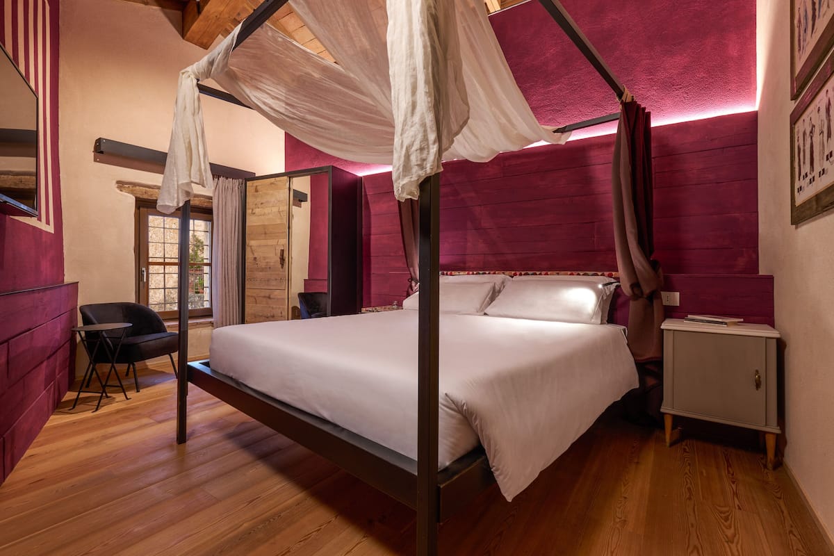 A four-poster bed with white linens, a wooden floor, a nightstand, and a chair.
