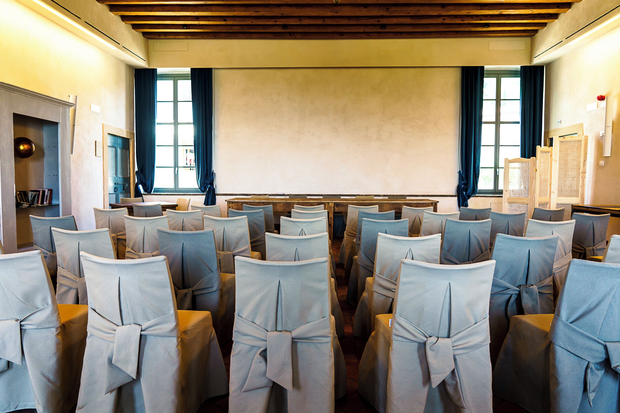 A conference room with rows of chairs covered in light blue fabric, wooden paneling on the ceiling, and large windows with curtains.