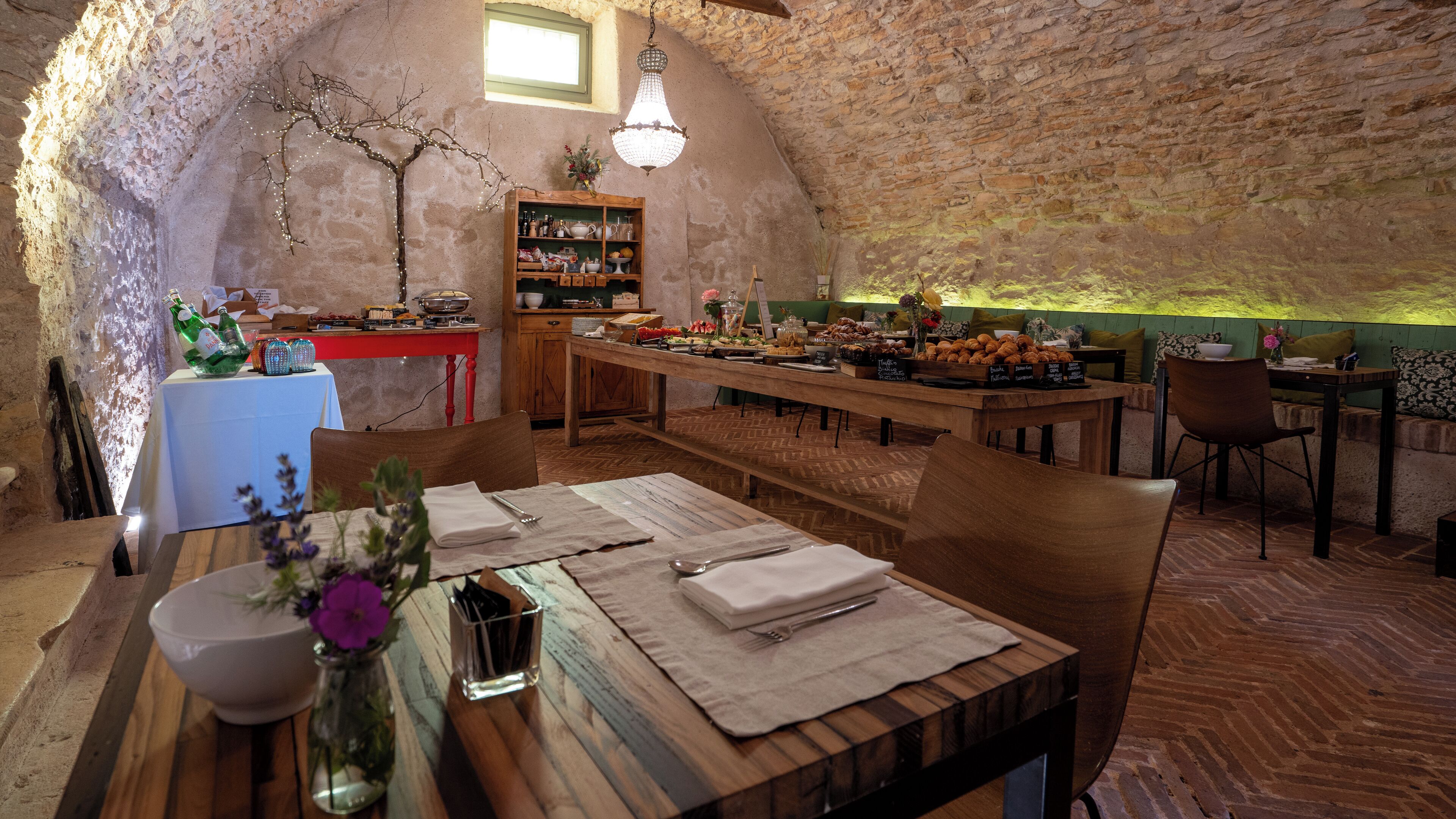 A rustic dining area with a wooden table set for two, a small vase with flowers, and a shelf with various items.
