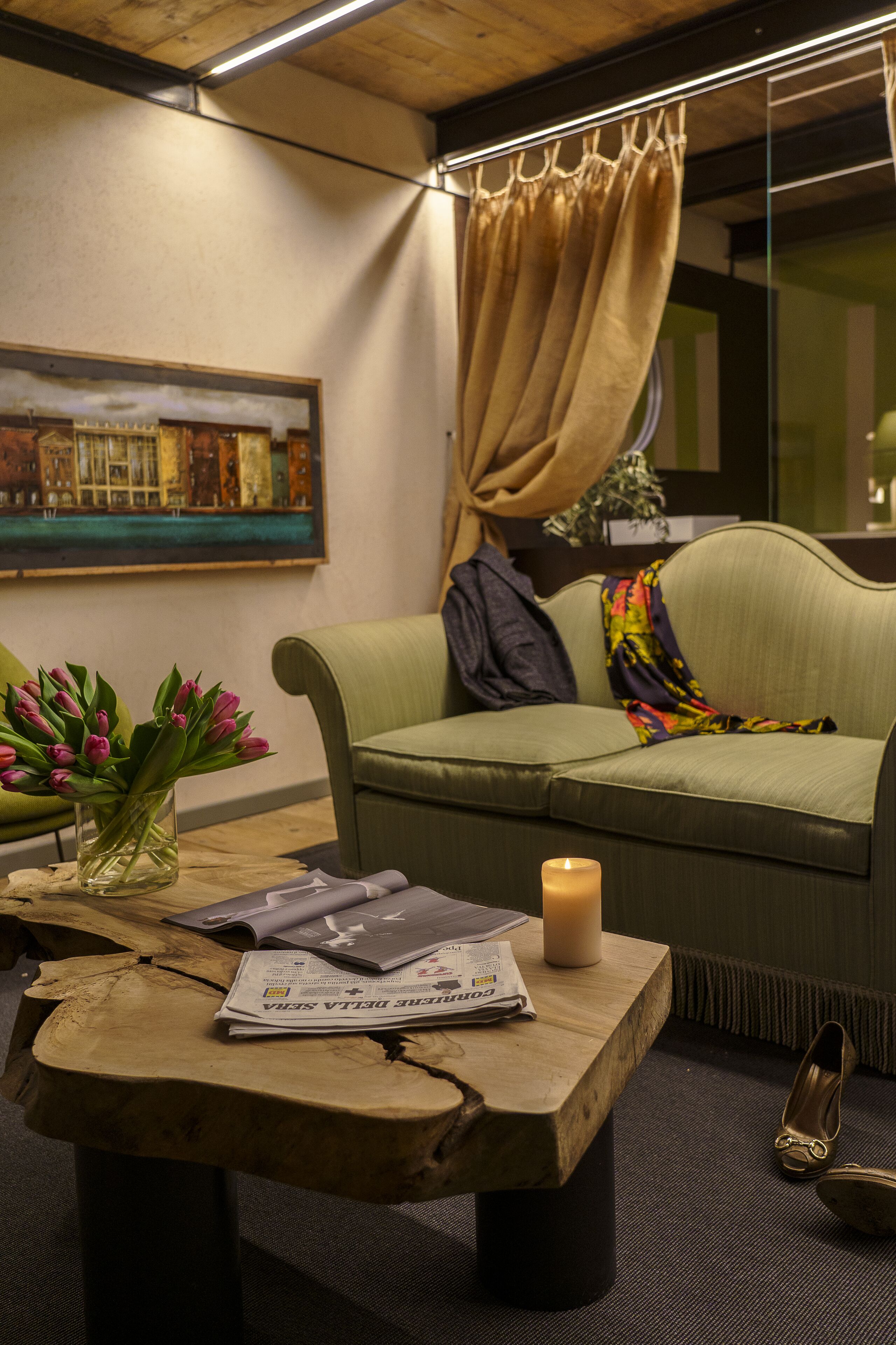 A cozy room with a green sofa, a wooden table with a newspaper, a vase of pink flowers, and a candle.