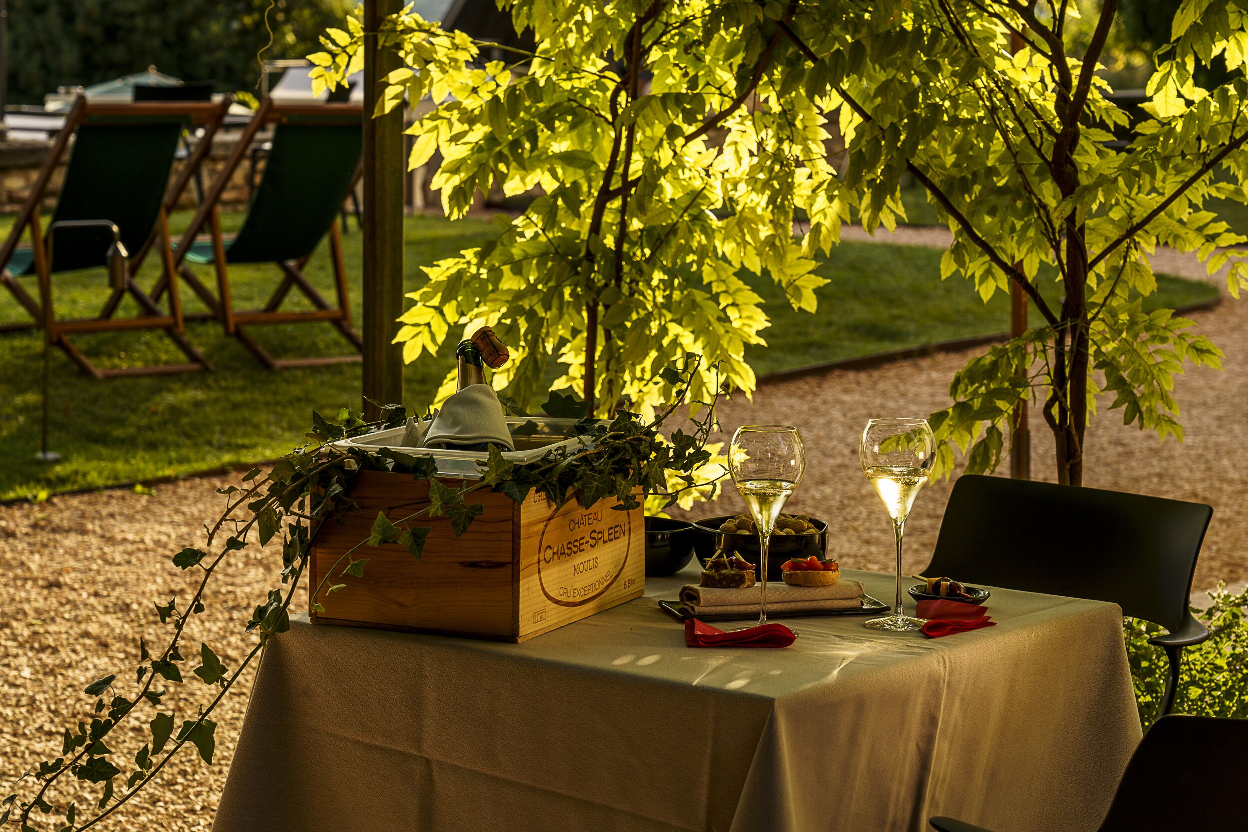 Un área de comedor al aire libre con una mesa dispuesta para dos, que incluye una caja de madera con una botella, una copa de vino blanco y un plato de comida.