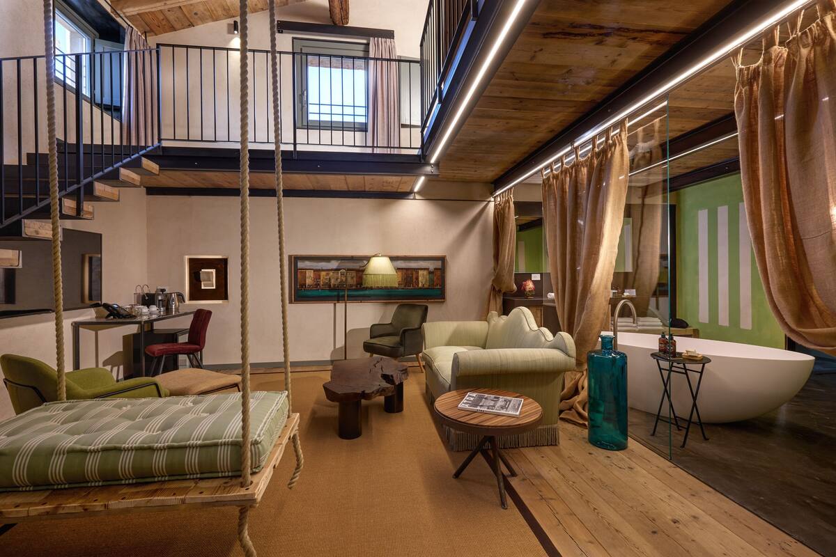A spacious living area with a high ceiling, a suspended bed, a dining table, and a large window with curtains.
