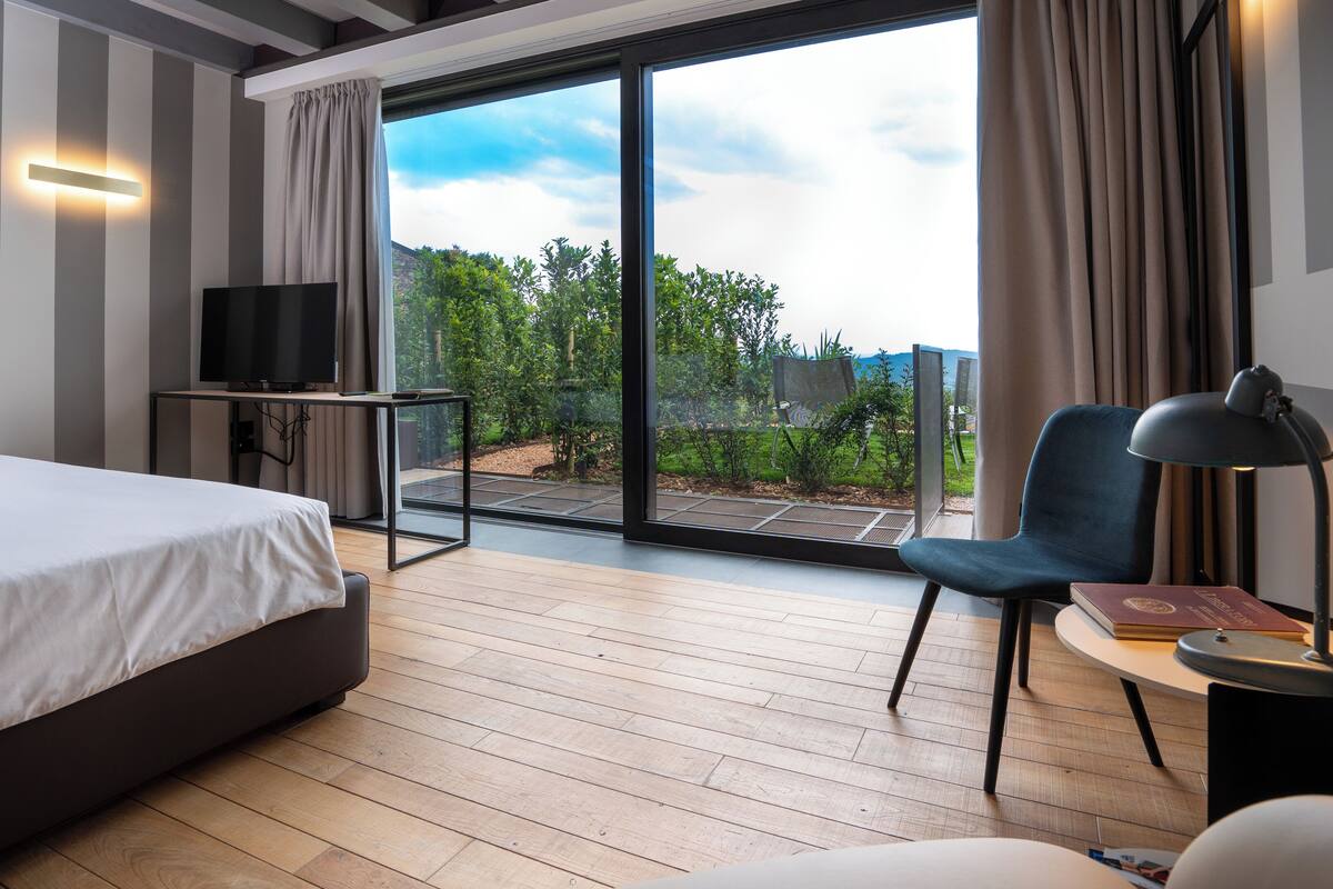 A modern hotel room with a large bed, a desk with a flat-screen TV, a chair, a lamp, and a view of a garden and mountains through the glass door.