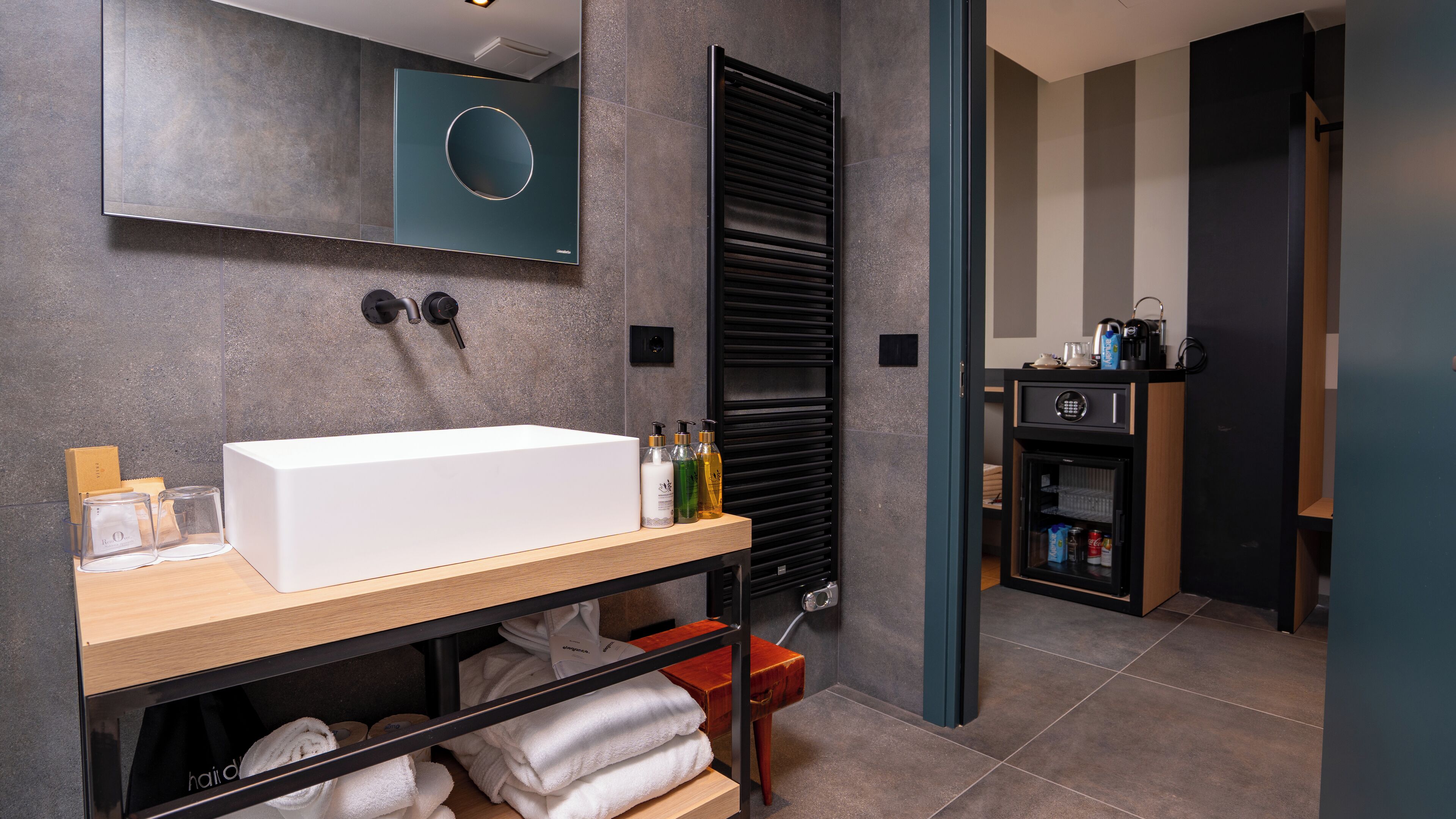 A modern bathroom with a large white sink, a mirror, a towel rack, and a built-in cabinet with toiletries.