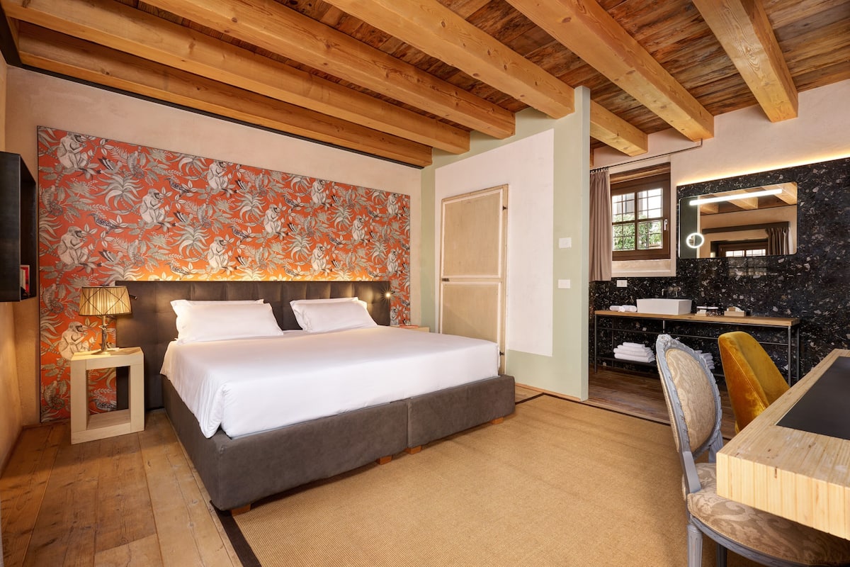A bedroom with a bed, a nightstand, a desk with a chair, a mirror, and a bathroom visible through an open door.