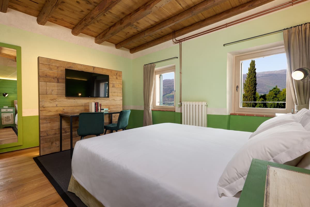 A hotel room with a wooden ceiling, a bed with white linens, a desk with a chair, a television mounted on a wooden panel wall, and a window with a view of the outdoors.