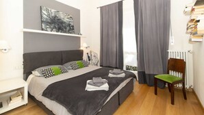 A bedroom with a bed, a chair, a radiator, and a shelf with books.