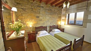 Una habitación con dos camas de madera, paredes de piedra, un aparador de madera, un espejo y una ventana con vistas al mar.