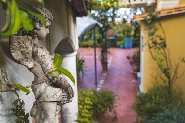 A statue of a winged figure on a column, a red pathway, and a yellow building with greenery.