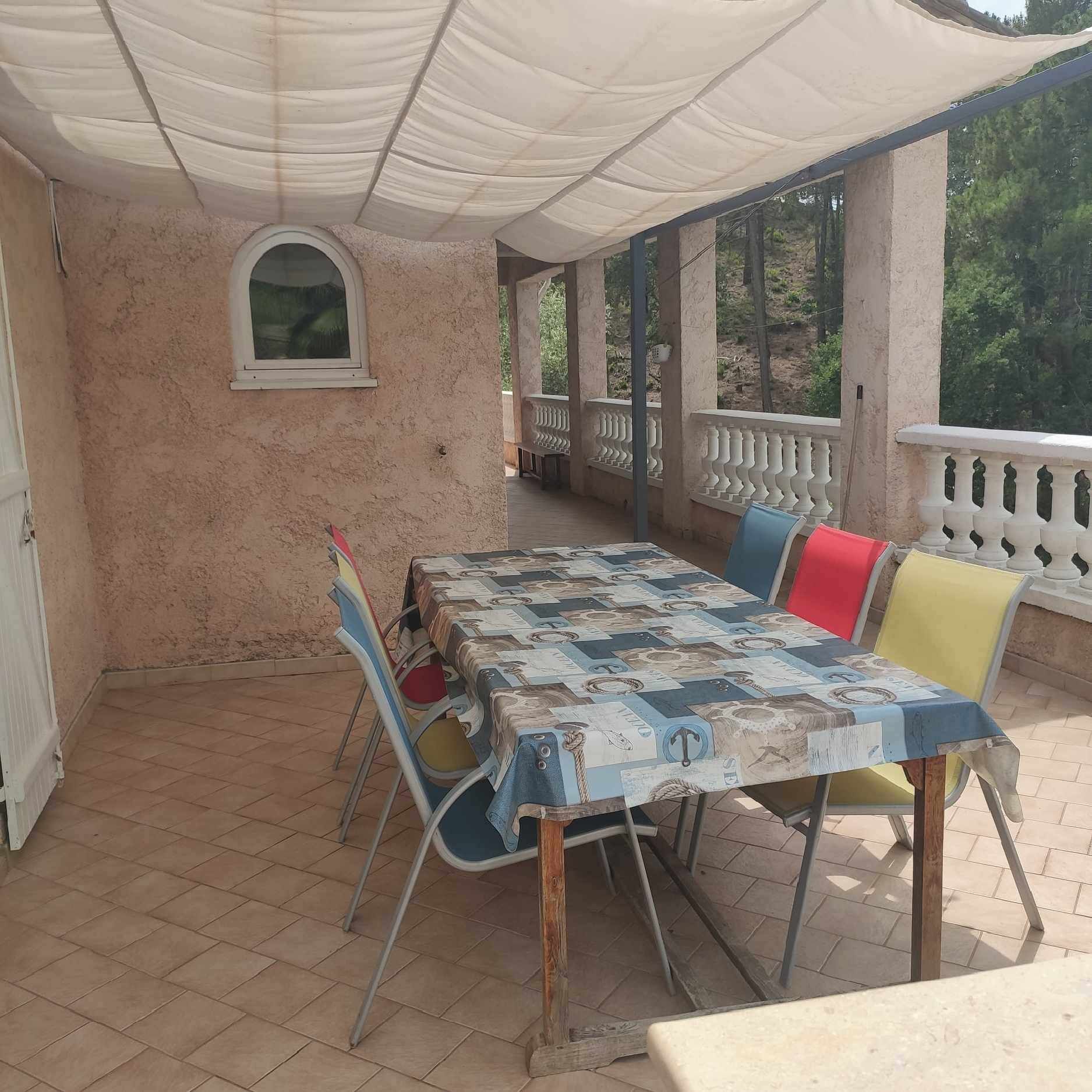 An outdoor patio with a table covered in a patterned tablecloth, surrounded by metal chairs with colorful cushions.