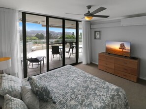 Une chambre à coucher avec un lit, un téléviseur posé sur une commode, un ventilateur de plafond et une porte-fenêtre coulissante donnant sur un balcon équipé de chaises et d’une table.