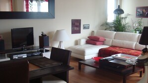 Een woonkamer met een flatscreen televisie, een bank, een salontafel en een eethoek met een stoel en tafel.