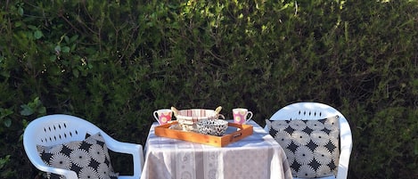 A small outdoor table with a tablecloth, two white plastic chairs with patterned cushions, and a tray with cups and a bowl.