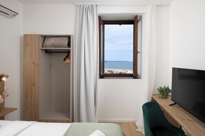 A bedroom with a bed, a TV, a window with a view of the beach, a wooden shelf, and a green chair.