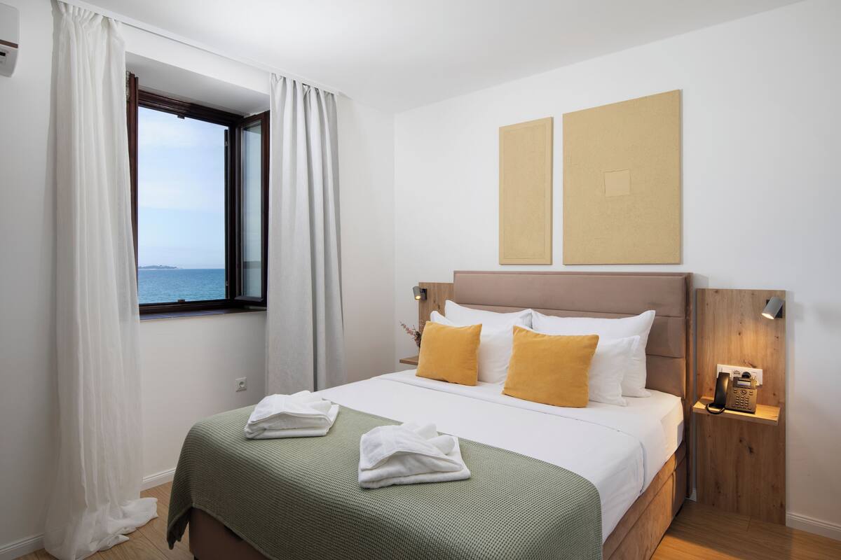 A hotel room with a bed, a window with curtains, a wall-mounted phone, and a view of the sea.