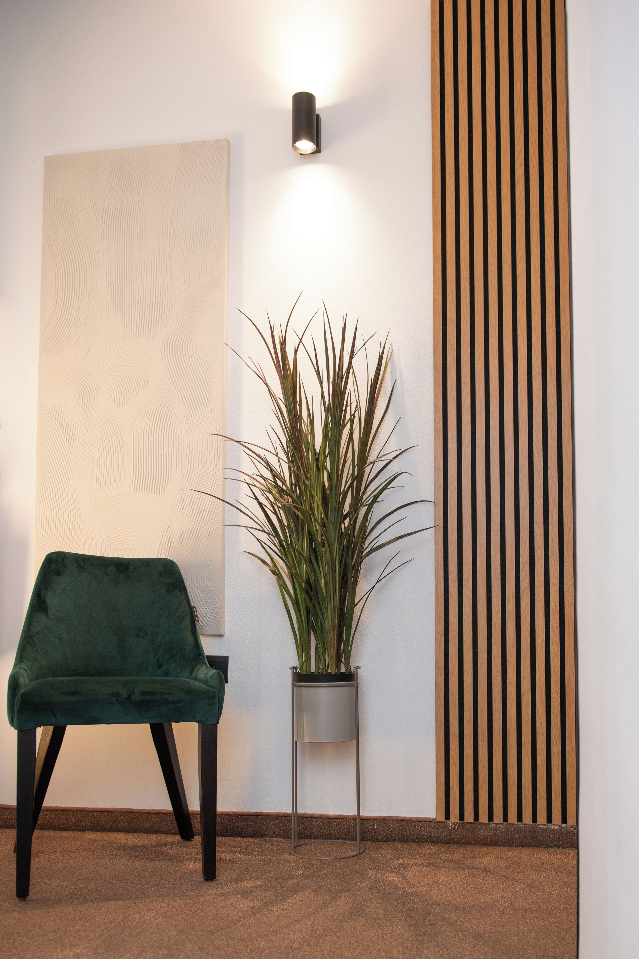 A room with a green velvet chair, a potted plant, a wall-mounted light, and a wooden slat panel.