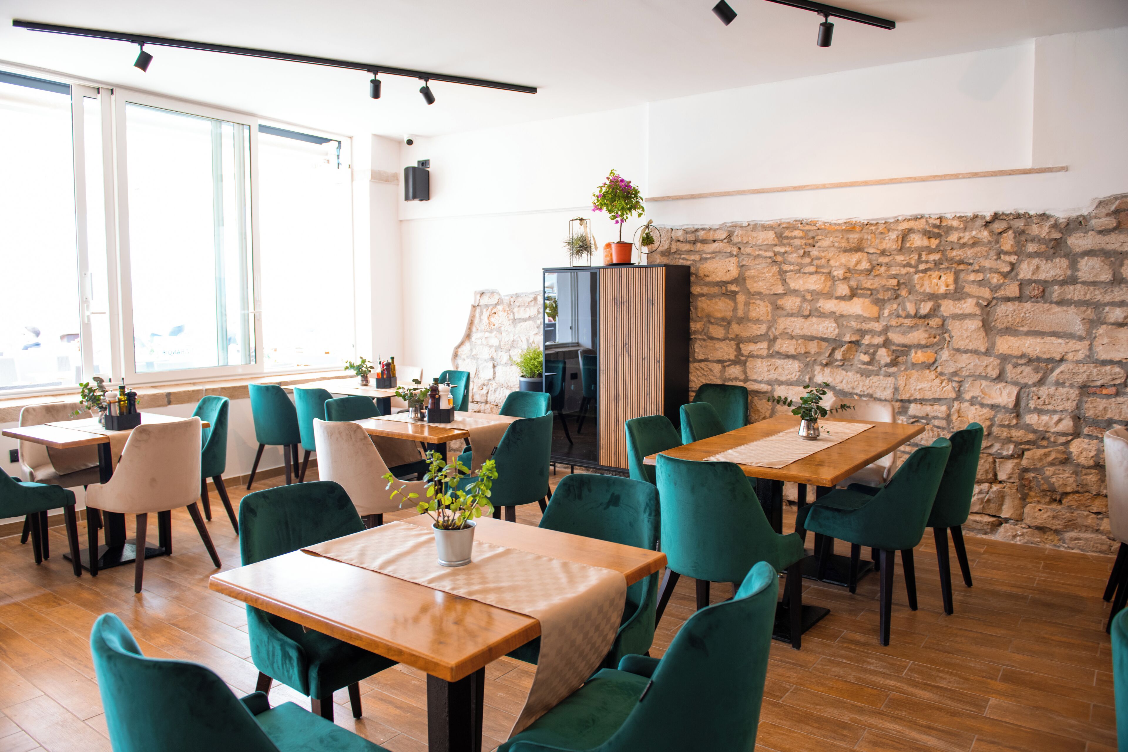 A restaurant with wooden tables and green chairs, a stone wall, and a large window.
