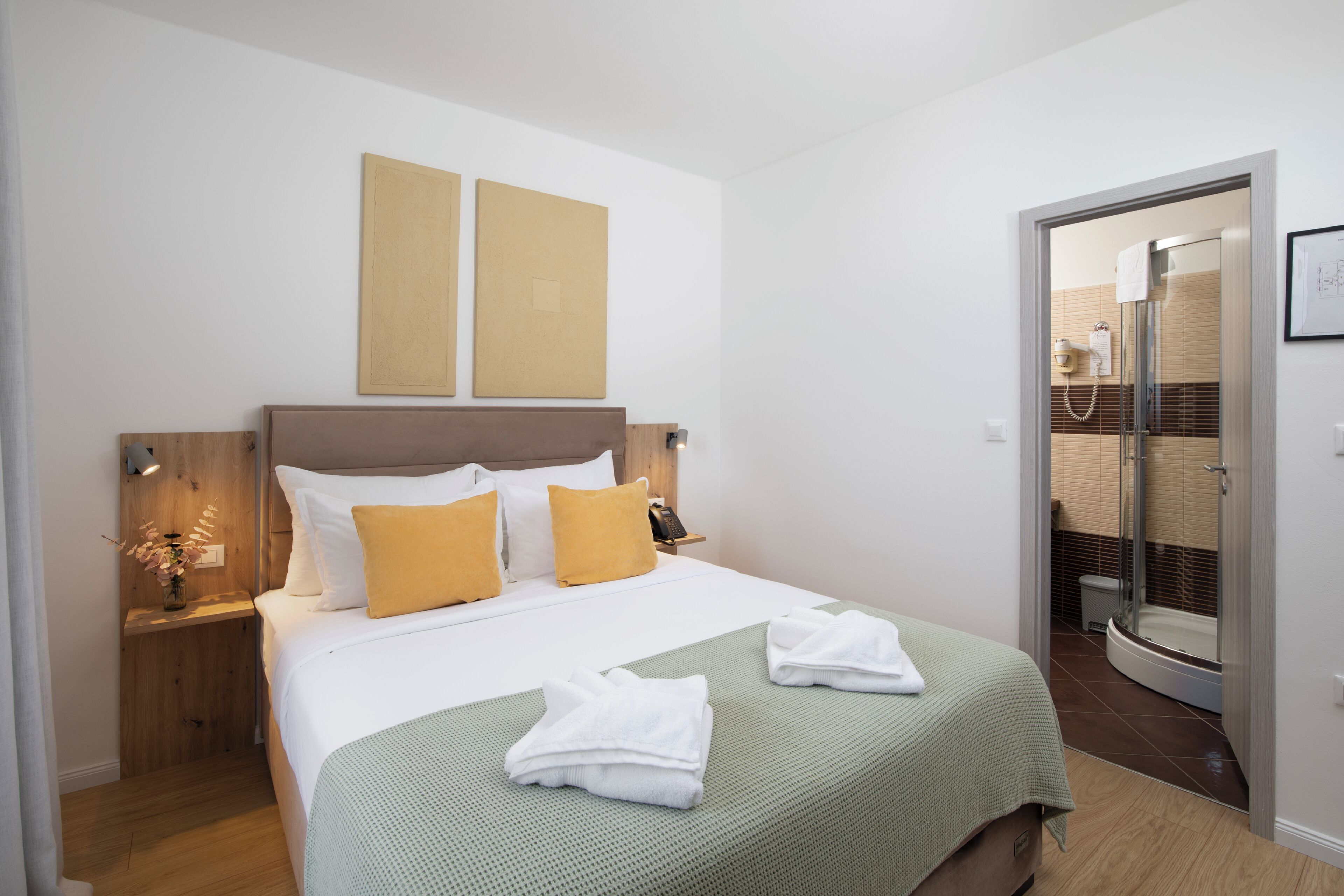 A hotel room with a bed, a nightstand, a bedside table with a lamp, a shower area, and a door leading to another room.