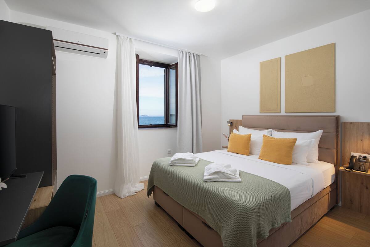 A hotel room with a bed, a desk, a chair, a window with curtains, and a view of the sea.