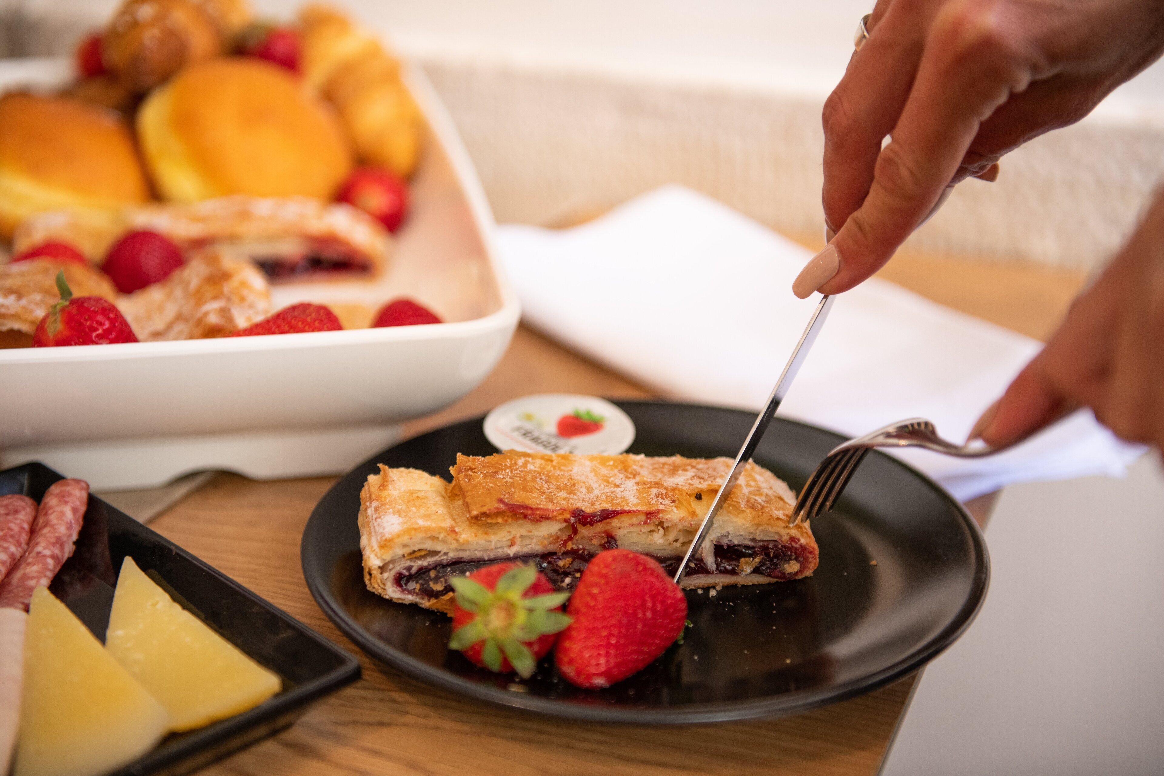A dessert plate with a pastry and strawberries, a tray of pastries, and a plate with cheese and sausages.
